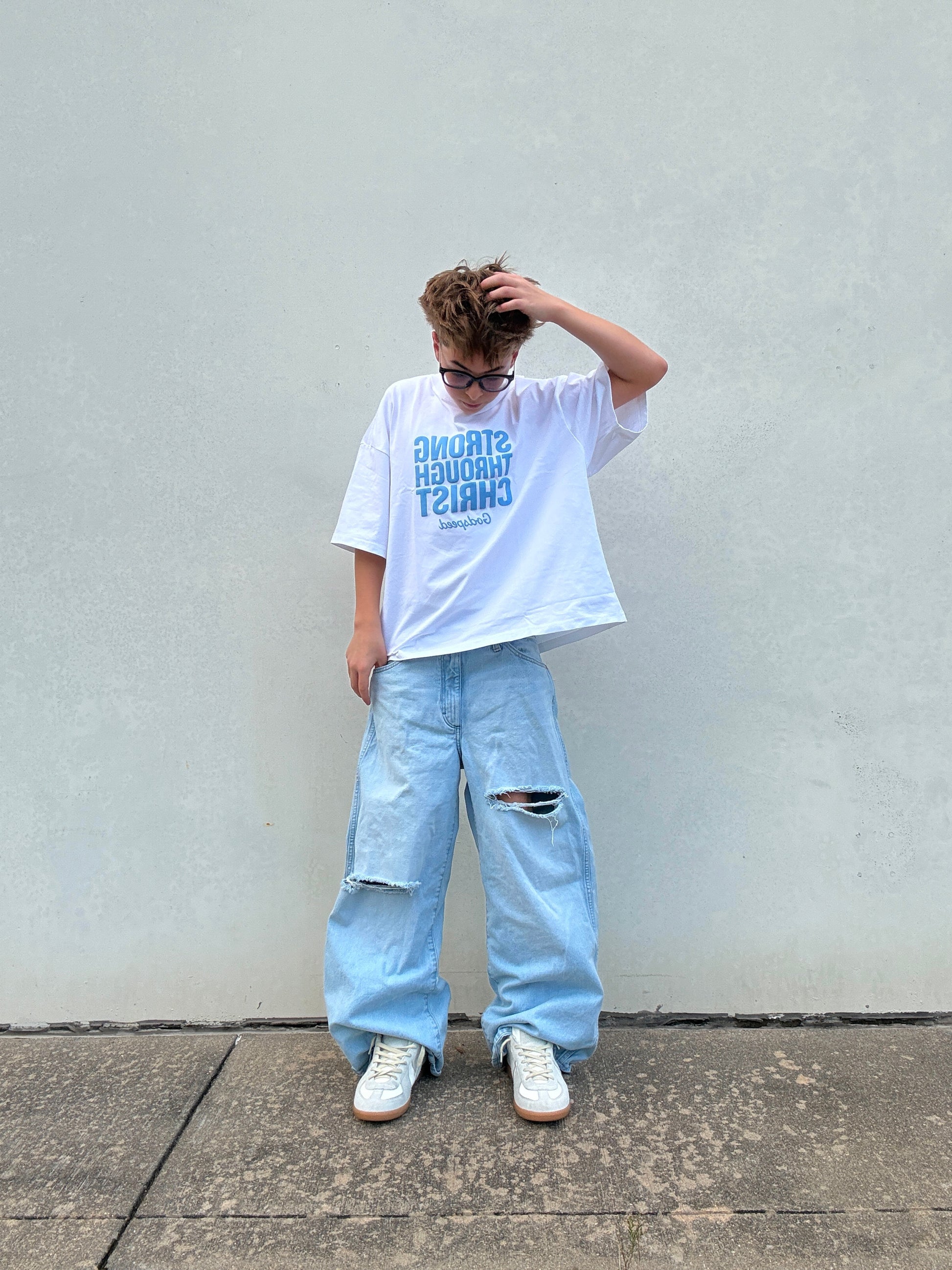 Person wearing a white t-shirt with text and light blue jeans against a plain wall.