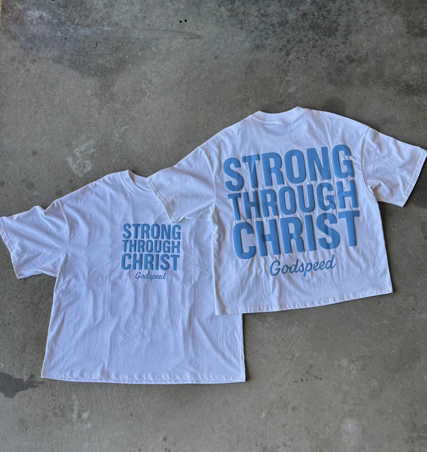 Two white t-shirts with blue text on a concrete surface